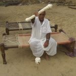 Sitting on his own work. These beautifully patterned chapoy are made for visitors to sleep on. The wooden frame is made by a carpenter.